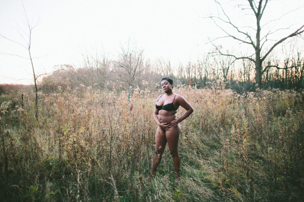 A wide angle shot of a Black nonbinary person wearing a lacey black demi cut bra and matching thong. They stand confidently, their stance widened and their hands on their right hip connecting together. The field is barely lit and the sun is down.